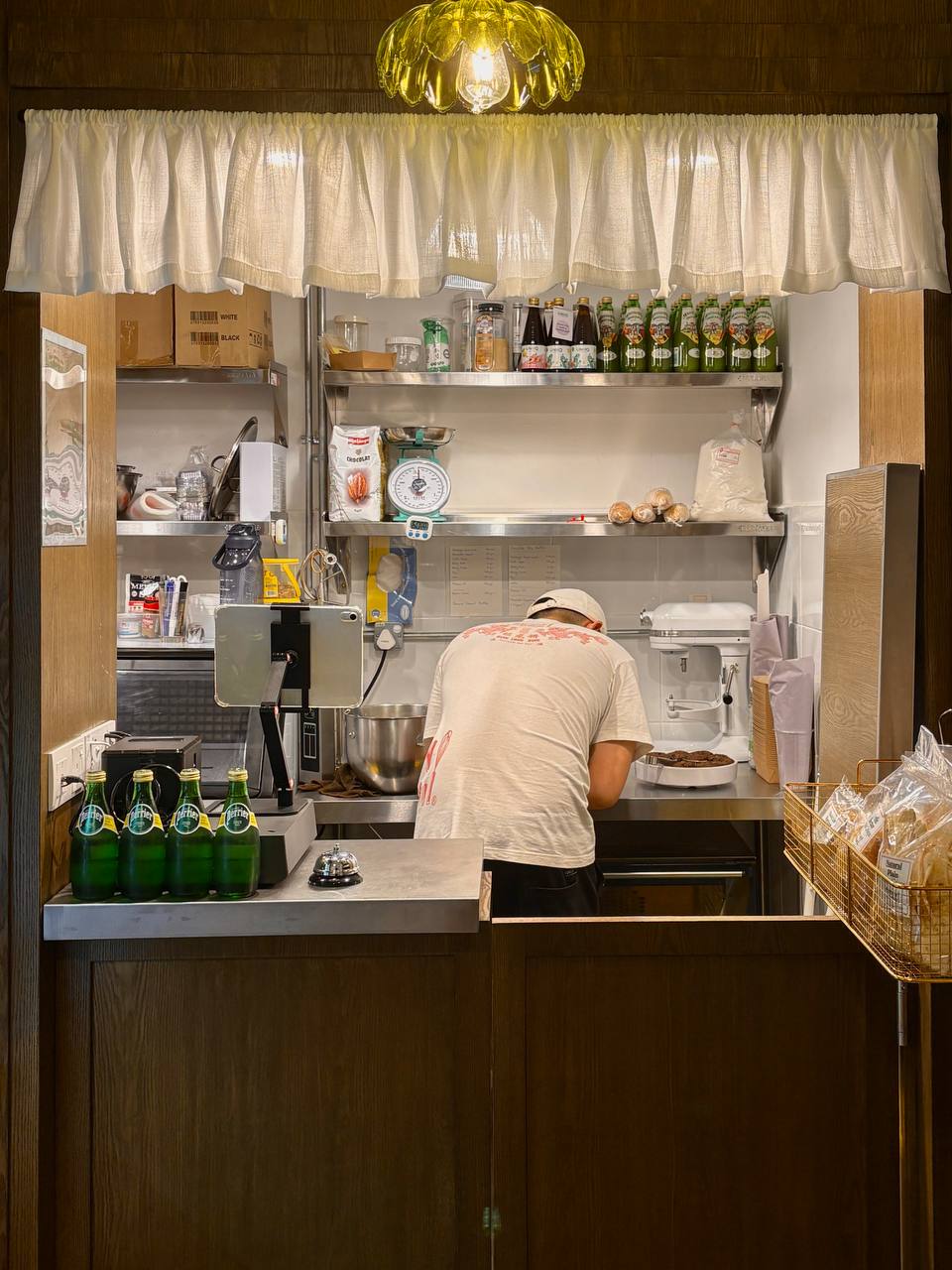 Tommy Le Baker service counter and stainless-steel workspace framed by café curtains and a pendant light